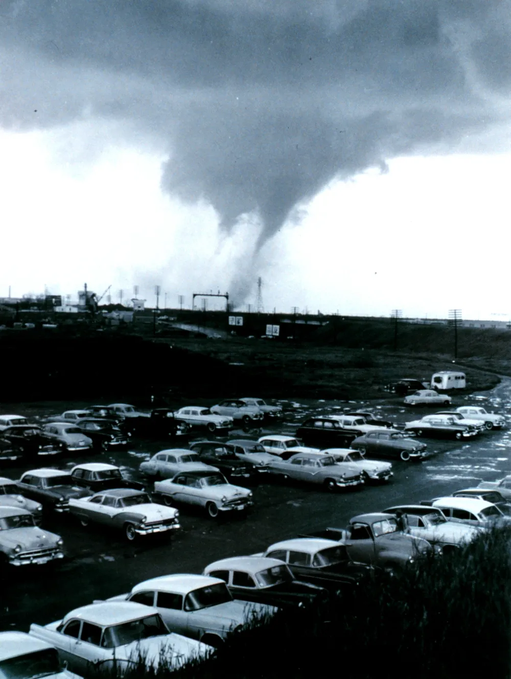 A black and white photograph of a tornado over a city with two distinct funnels side by side beneath the cloud base, seen across a parking lot full of 1950s cars.
