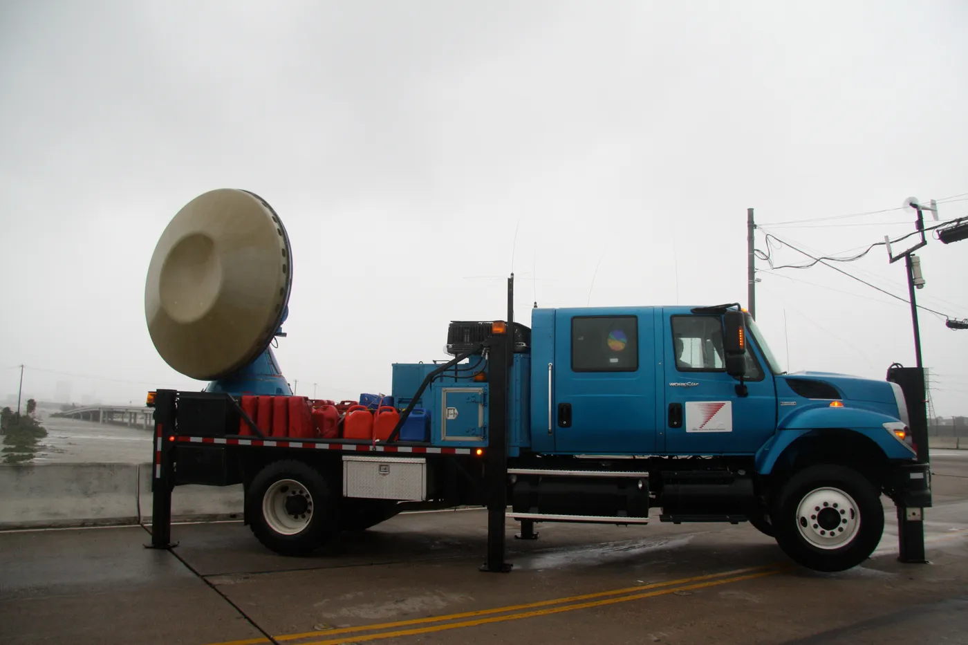 A blue flatbed truck carrying a large radar dish, parked on a wet road under a gray sky with power lines behind.