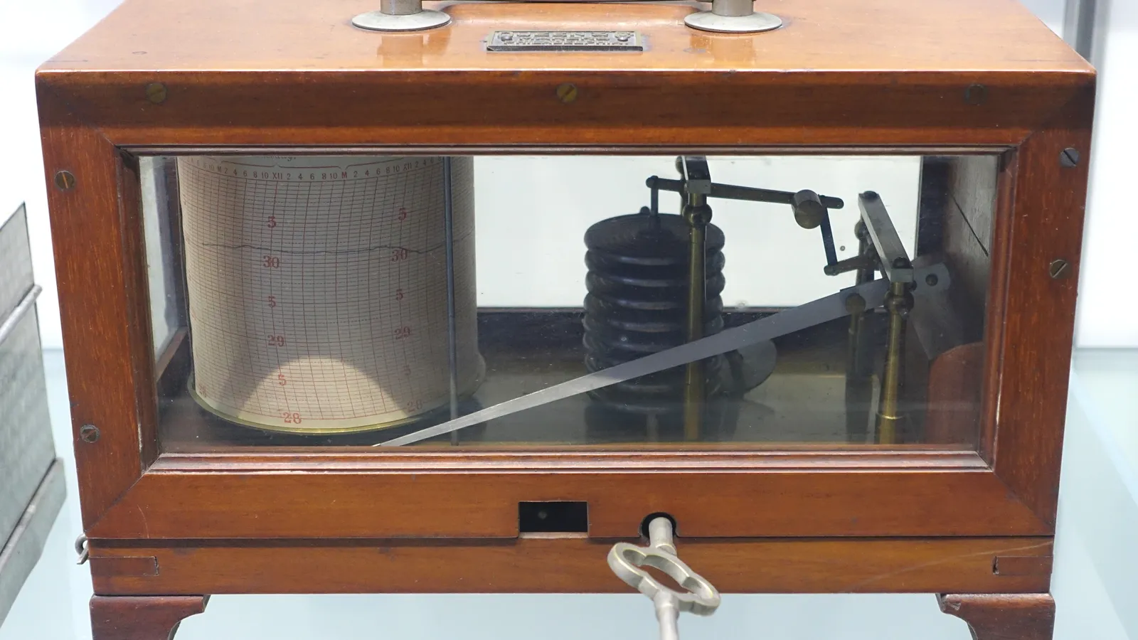 A recording barograph in a wooden case with a glass front: a paper chart wrapped on a drum at left with a faint ink trace near 30, and at right a stack of dark aneroid capsules linked by levers to a long pen arm.
