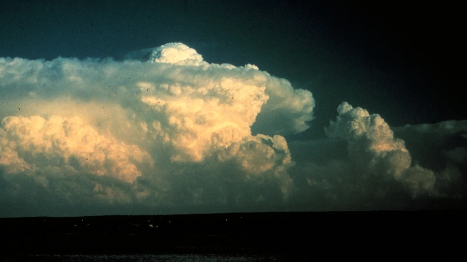 Towering cumulonimbus clouds lit gold by a low sun against a dark sky, with a rounded dome of cloud pushing up above the flat top of the largest one and a younger cumulus tower to its right.