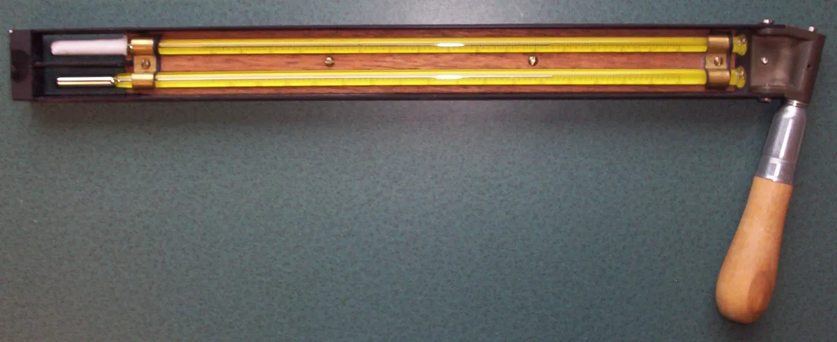 A sling psychrometer: two long glass thermometers in a wooden and metal frame, the upper one with a white muslin sleeve over its bulb at the left end, and a wooden handle at the right.