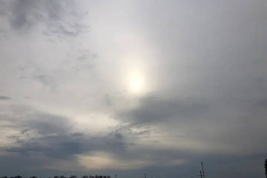The sun seen as a bright, softened disk through a thin whitish veil of cloud covering most of the sky, over flat land with buildings and power lines on the horizon.