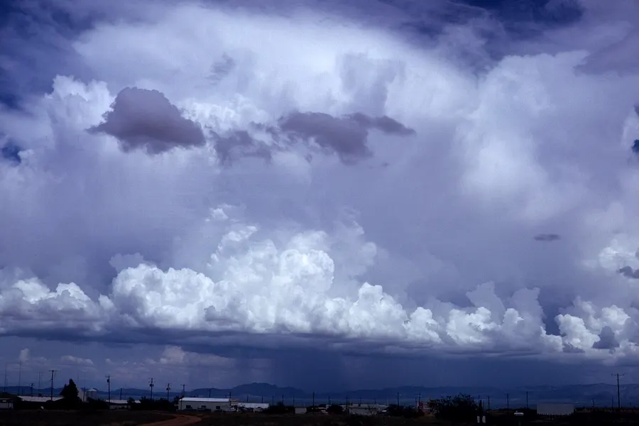 A towering storm cloud over flat desert with mountains on the horizon: a row of bright cumulus towers below and a broad, smooth upper mass spreading above, with rain falling beneath.