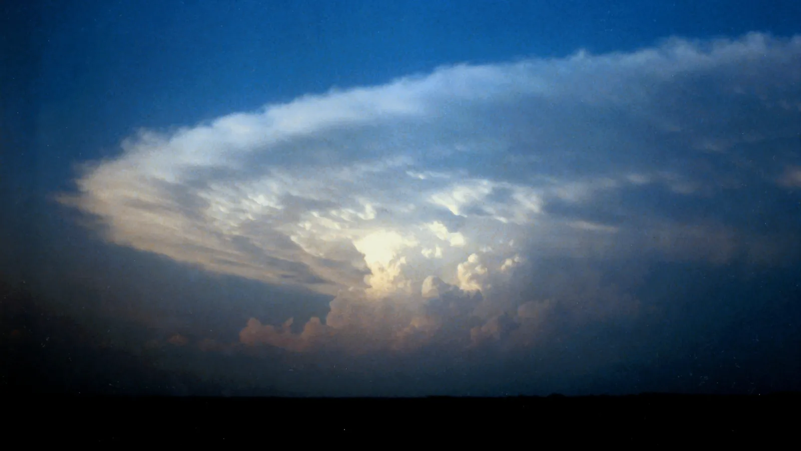 A thunderstorm seen from a distance at dusk over flat, dark ground: a broad white anvil fans out across a deep blue sky above a lit core of billowing towers.