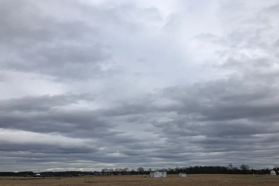 Long gray rolls and patches of cloud with darker undersides stretching across the sky over a flat field and a small white farmhouse.