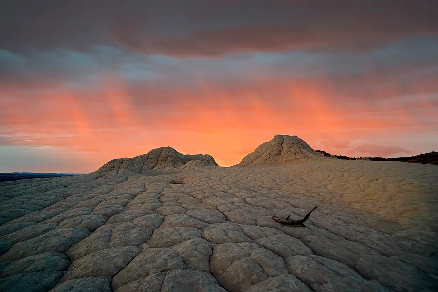 Streaks of precipitation hanging from clouds lit orange and pink at sunset, fading out above pale, dome-shaped sandstone formations.