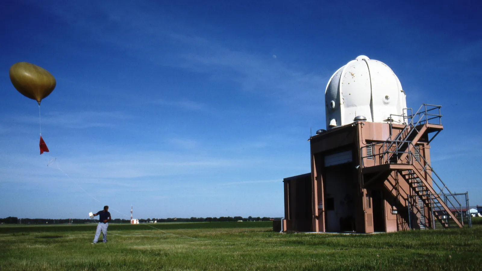 A meteorologist in a field holds a radiosonde while its balloon and a red parachute rise ahead of it on a line, beside a domed building at the Topeka, Kansas weather office.