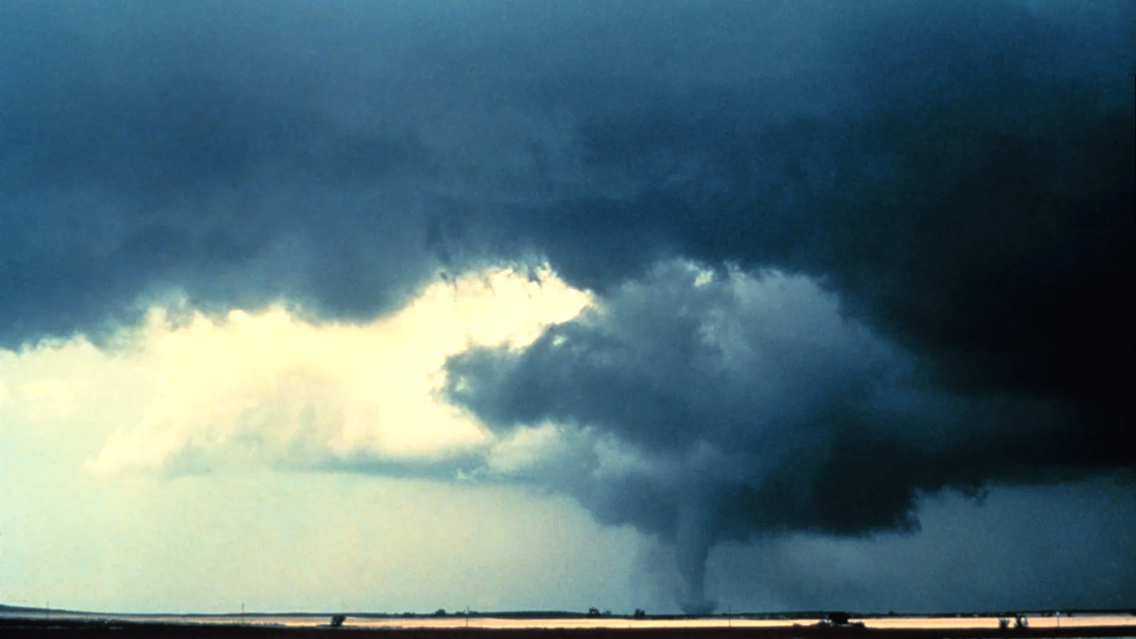 A tornado hangs from the dark base of a supercell over flat farmland, with a bright gap of clear sky beside it where dry air is sinking.