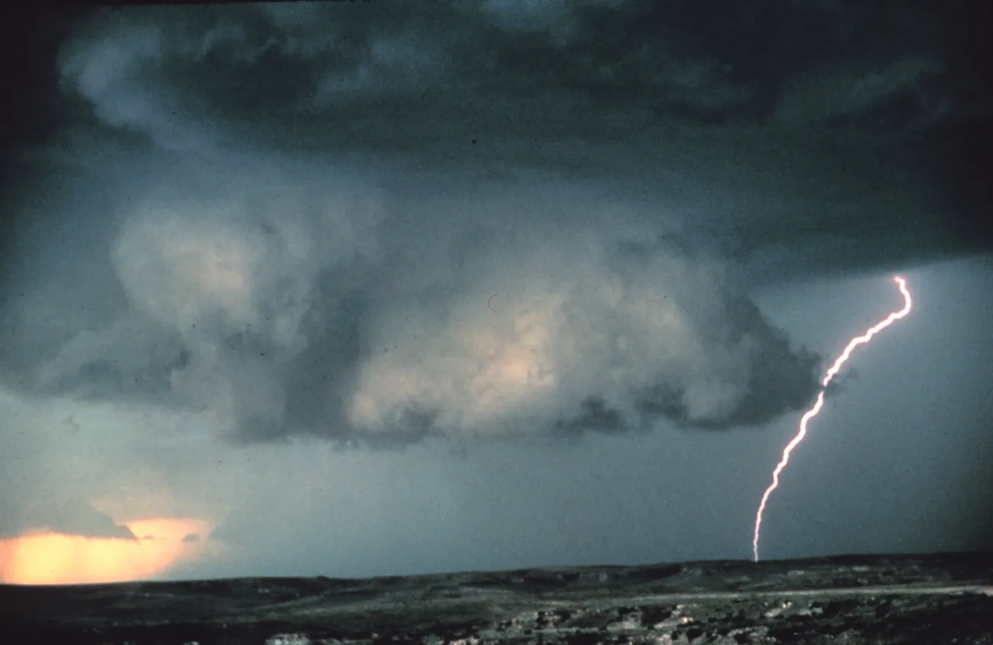 A broad, lowered block of cloud hangs beneath a dark storm base over rolling ground, with a lightning bolt striking to the right.