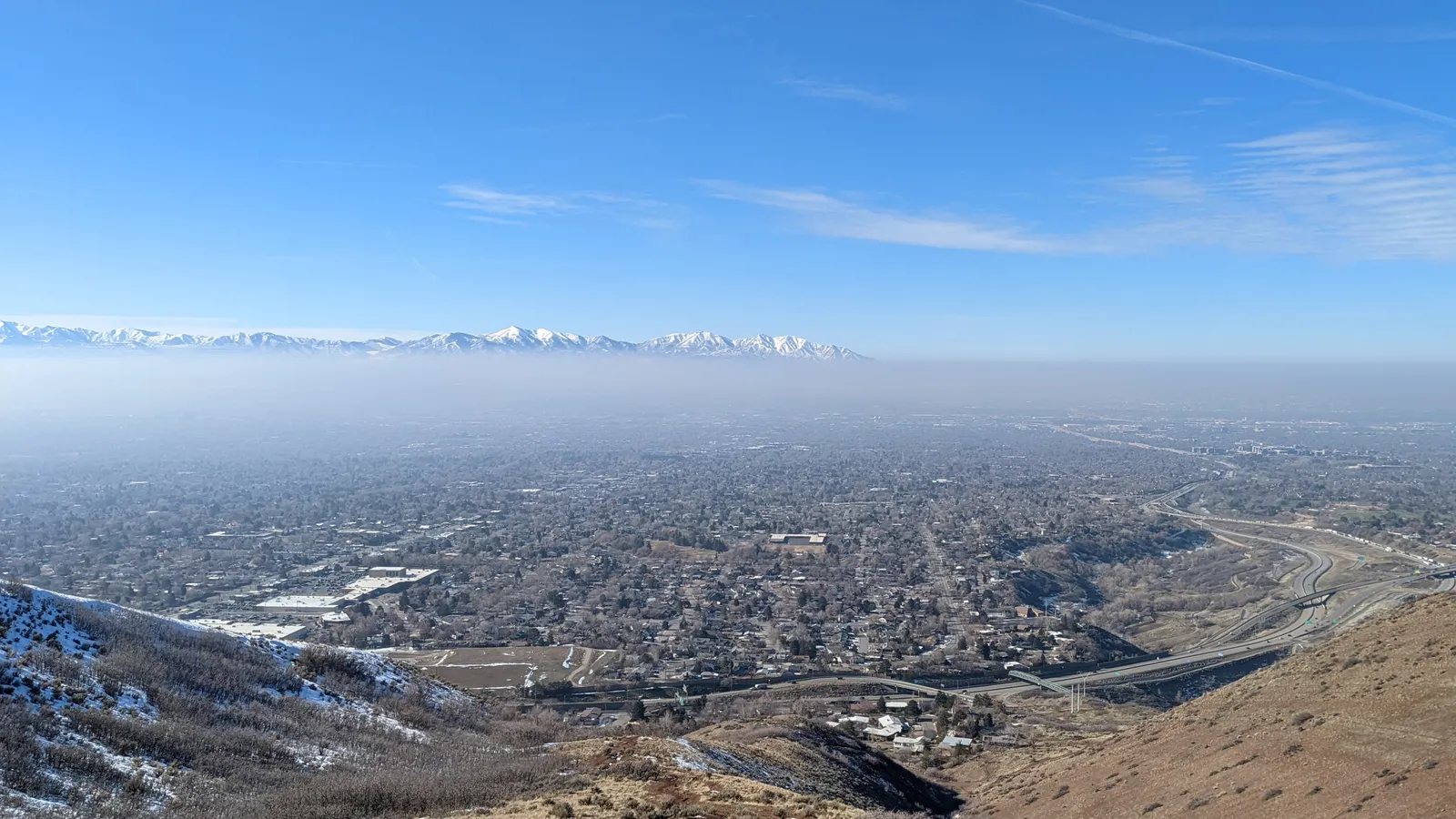 A wide view from a snowy hillside over the Salt Lake Valley: the city fills the valley floor under a flat layer of gray haze, and snow-capped mountains rise above the haze into a clear blue sky.
