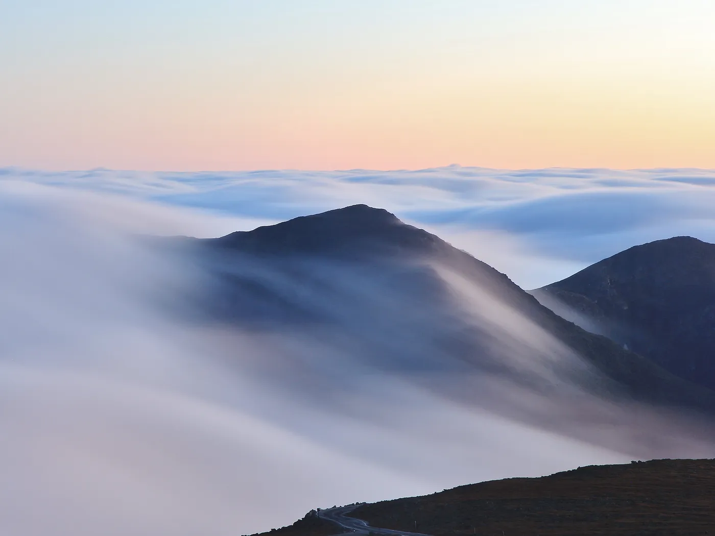 A dark mountain ridge at dawn with a smooth layer of cloud pouring over its shoulder like a waterfall, and a flat sea of cloud stretching to the horizon under a pale orange sky.