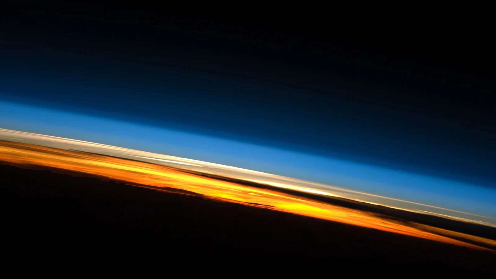 The edge of the earth at sunset seen from the International Space Station: a dark surface, a bright orange band of lower atmosphere, a thin pale layer above it, then light blue fading through deep blue into black space.