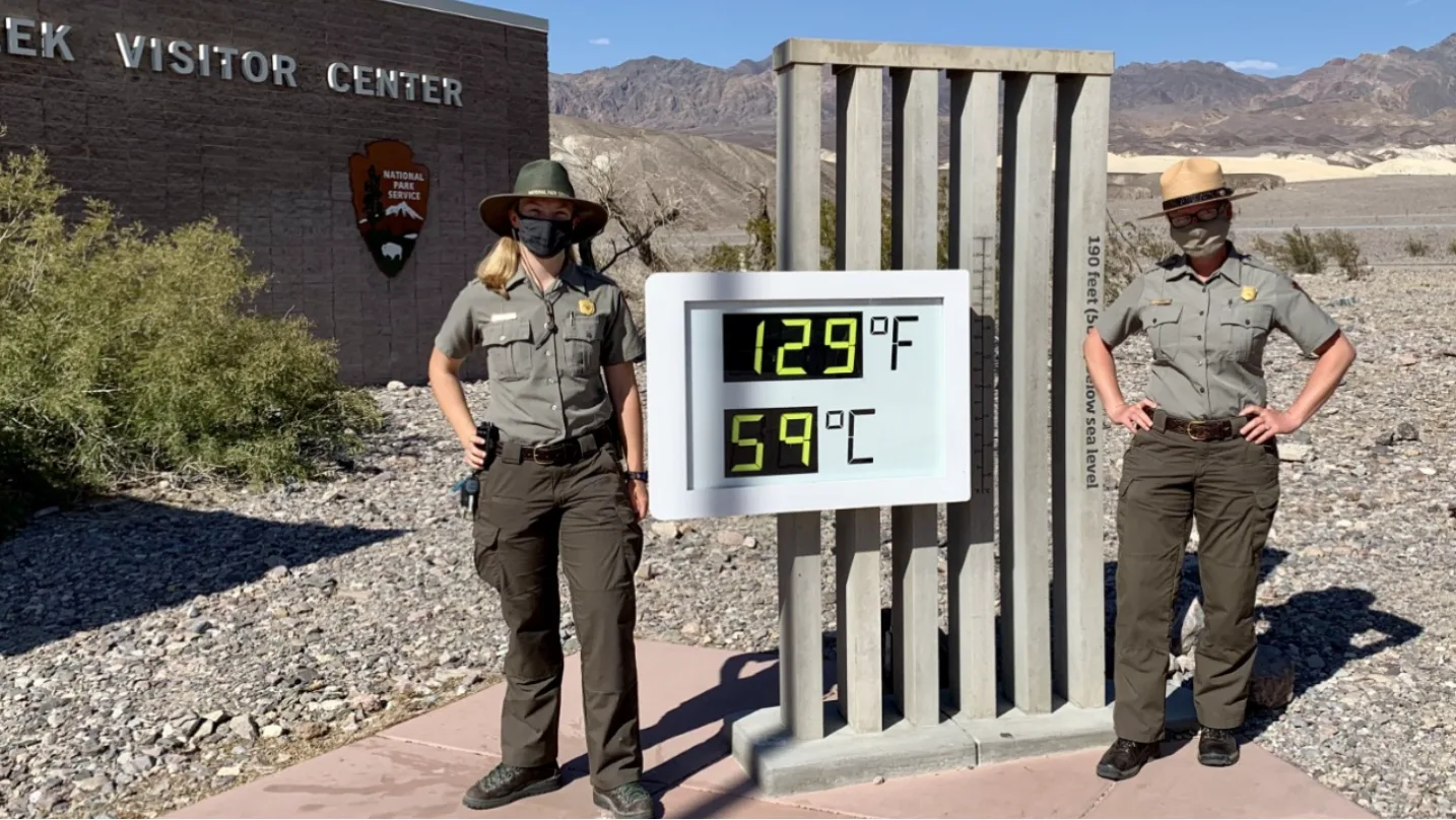 Two National Park Service rangers in masks stand on either side of a digital sign reading 129 degrees Fahrenheit and 59 degrees Celsius, outside the Furnace Creek Visitor Center in Death Valley.