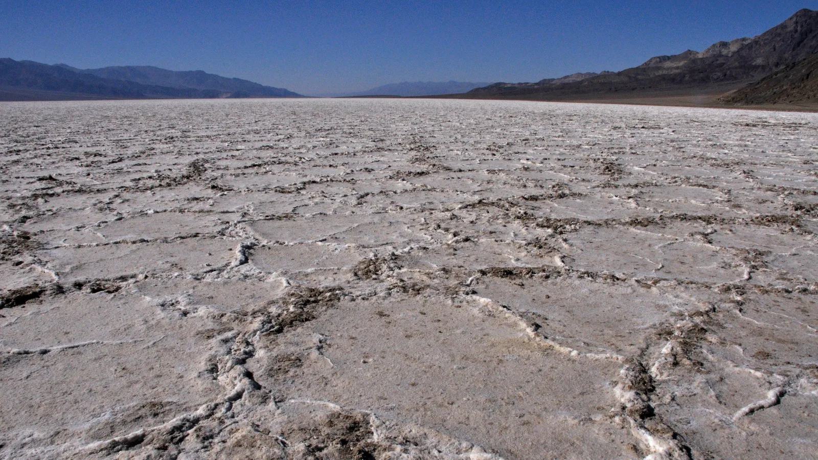 A white salt flat broken into polygons stretches to the horizon under a clear blue sky, with dark mountains on both sides of the valley.