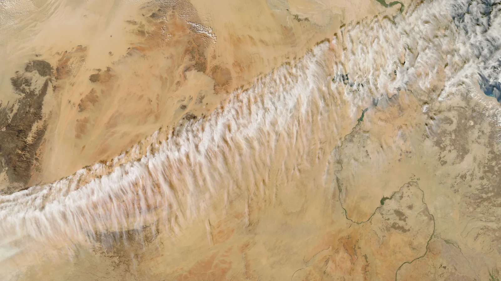 A satellite view of the Sahara in tan and orange, crossed diagonally from lower left to upper right by a long, broad band of white cirrus made of many short streaks lying across the band; the thin line of the Nile winds across the lower right.