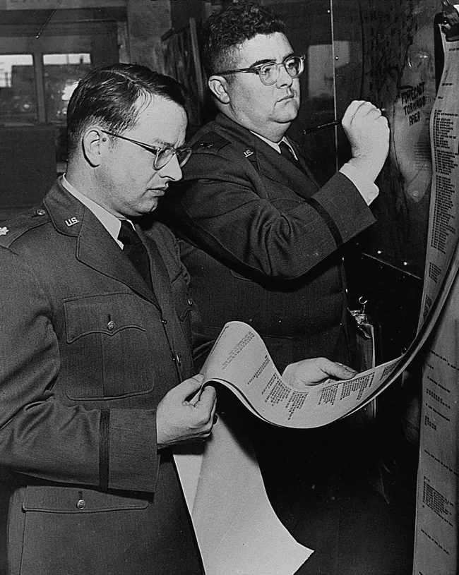 Two Air Force officers in uniform at a wall chart: Ernest Fawbush in the foreground reading a long teletype printout, Robert Miller behind him writing on a board headed forecast tornado area.