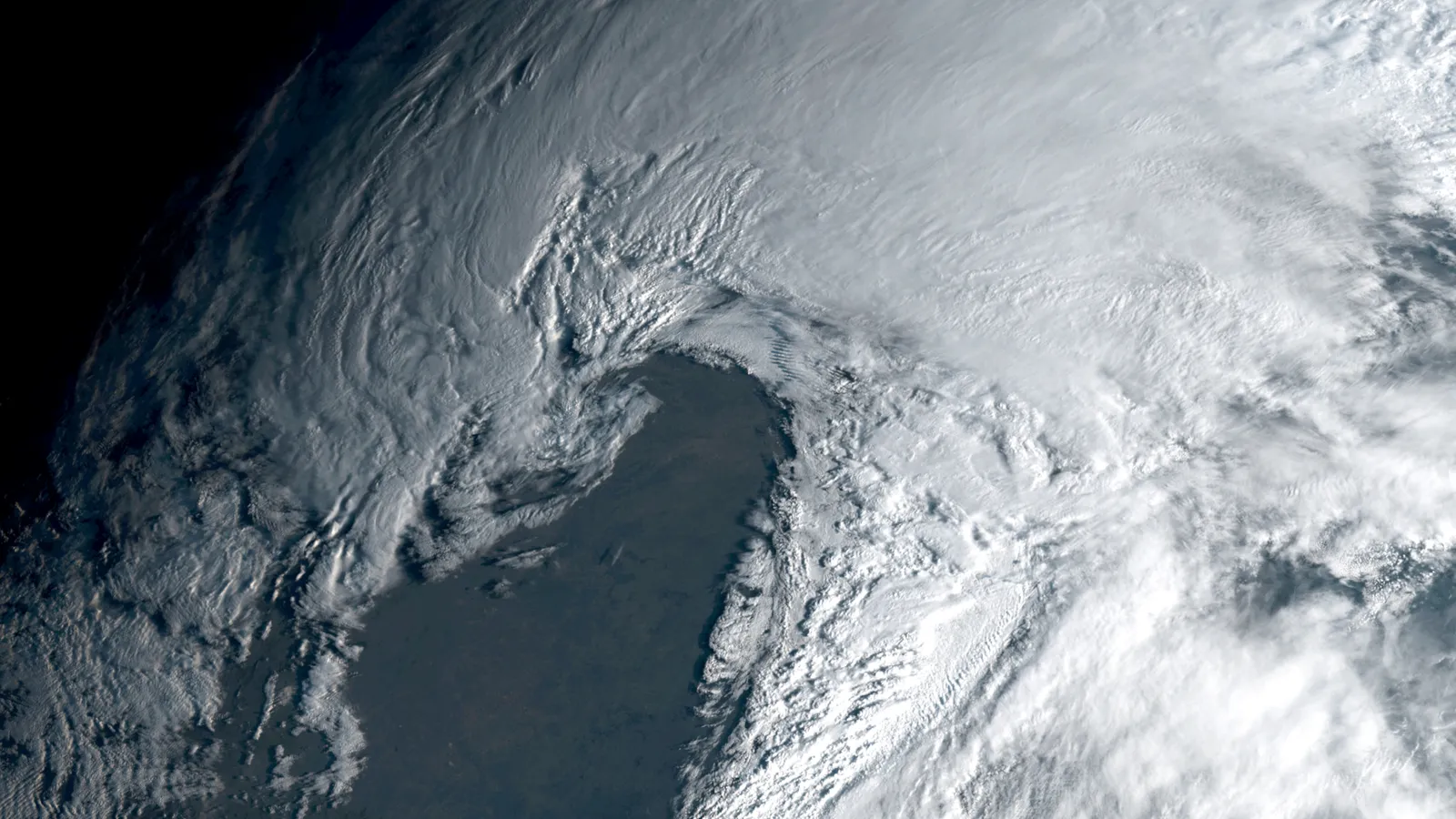 A satellite view of a large storm from above: a broad white cloud shield sweeps across the top and right of the picture and curls around a dark, cloud-free wedge in the center, where the ground shows through.