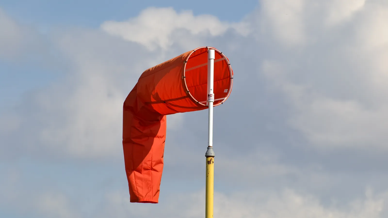 An orange airport windsock on a white and yellow pole against a blue sky with white clouds: the throat of the sock is filled with air and points into the wind, while its tail hangs down.