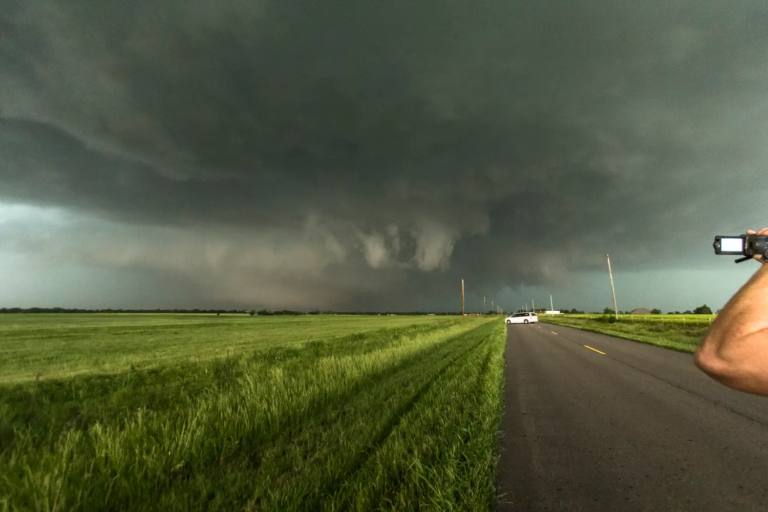 A vast dark rain-wrapped tornado filling the horizon beyond a green Oklahoma field, with a parked van on the road and a storm chaser's camera in the foreground.