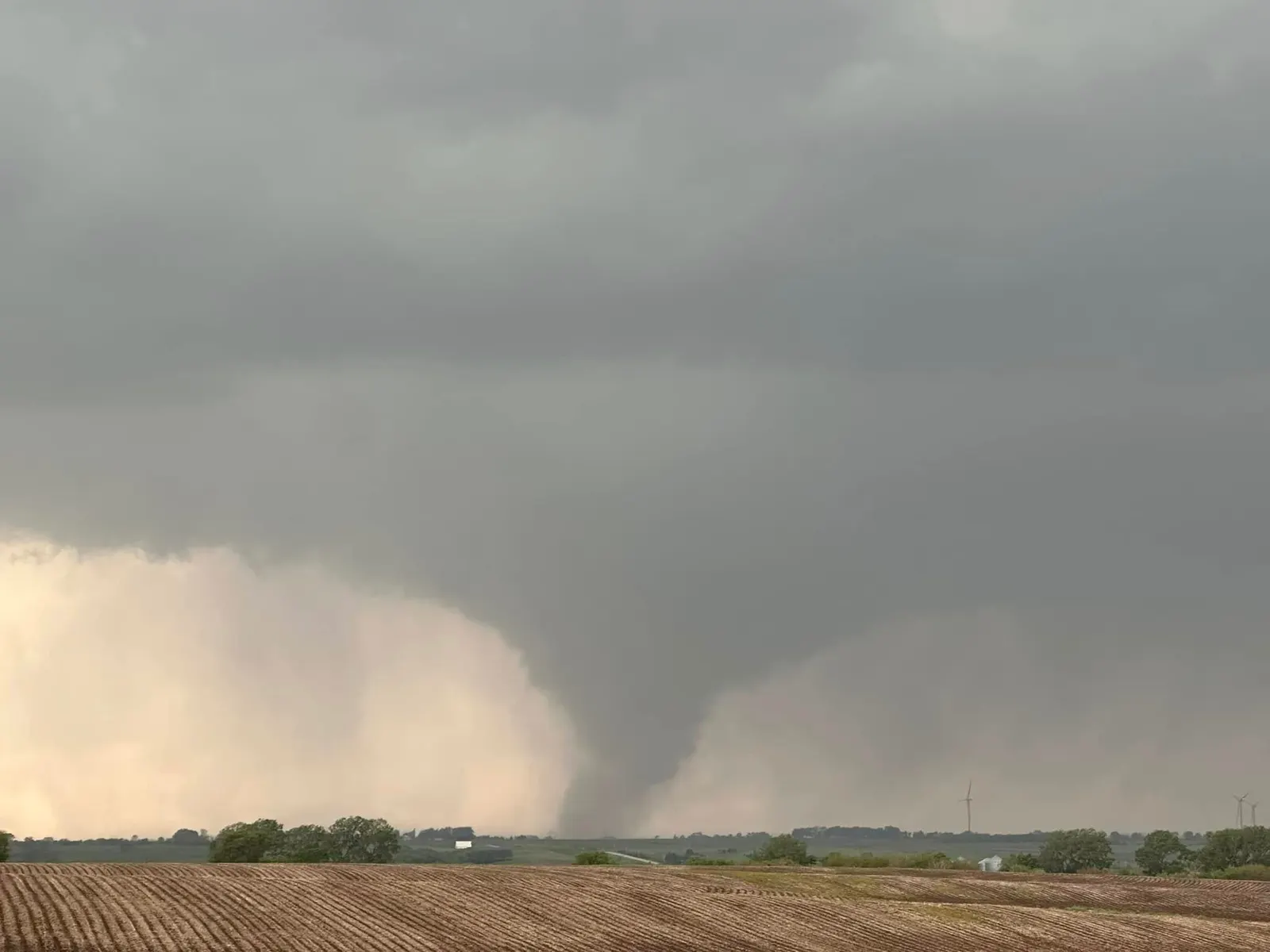A wide grey tornado on the horizon beyond a plowed Iowa field, with wind turbines visible to its right.