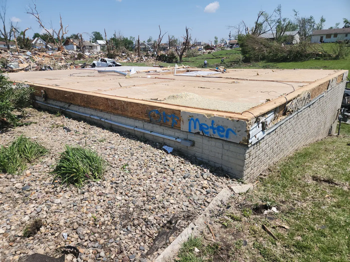 A bare foundation with only the subfloor remaining, spray-painted 'off meter', with rubble and shredded trees in the background.