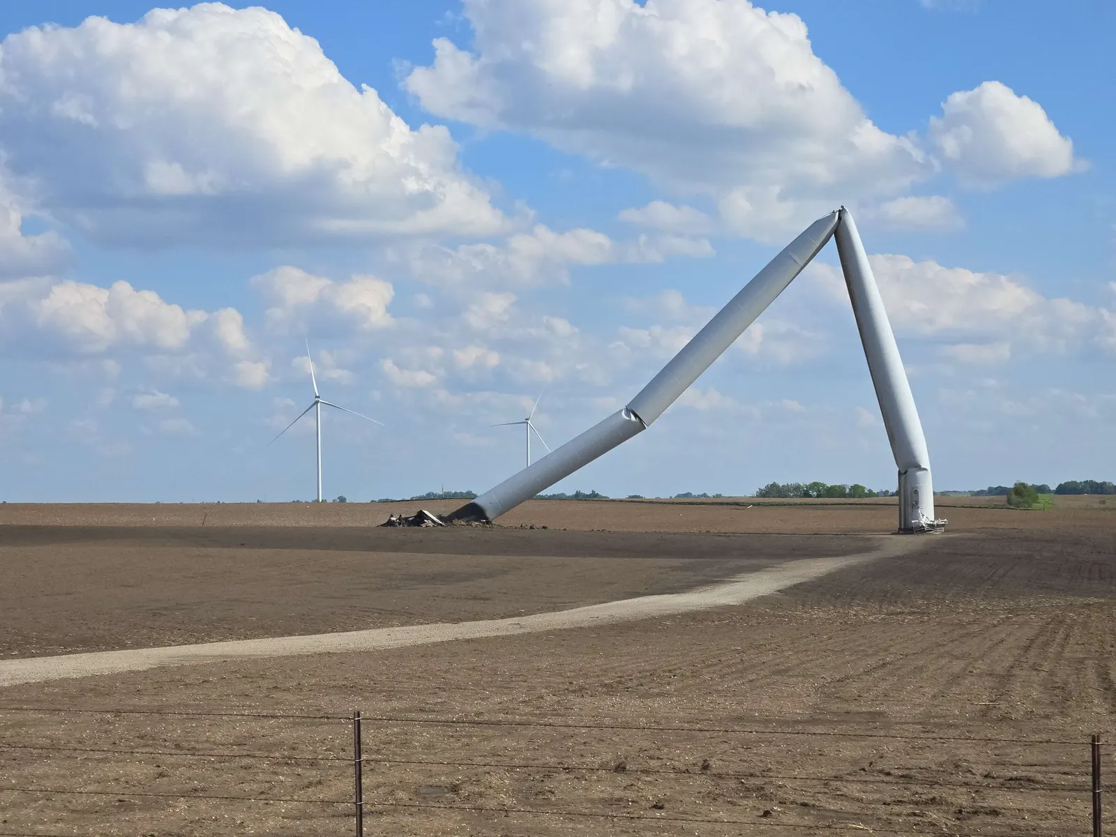 A utility-scale wind turbine folded to the ground in an open field, its tower buckled in two places, with intact turbines on the horizon.