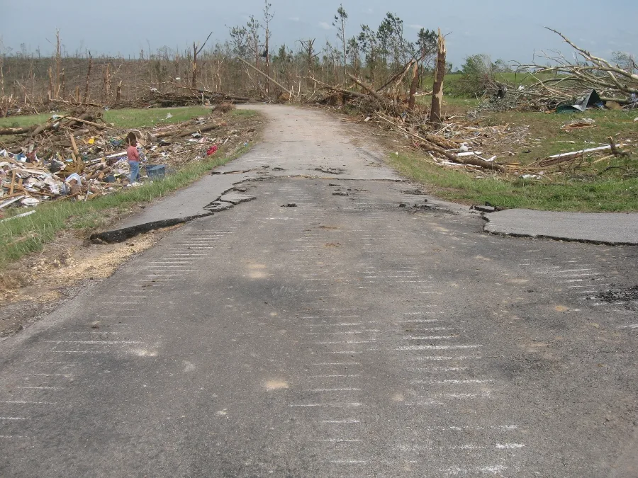 A rural road with its asphalt surface peeled away in patches, lined by snapped and debarked trees and piles of house debris.