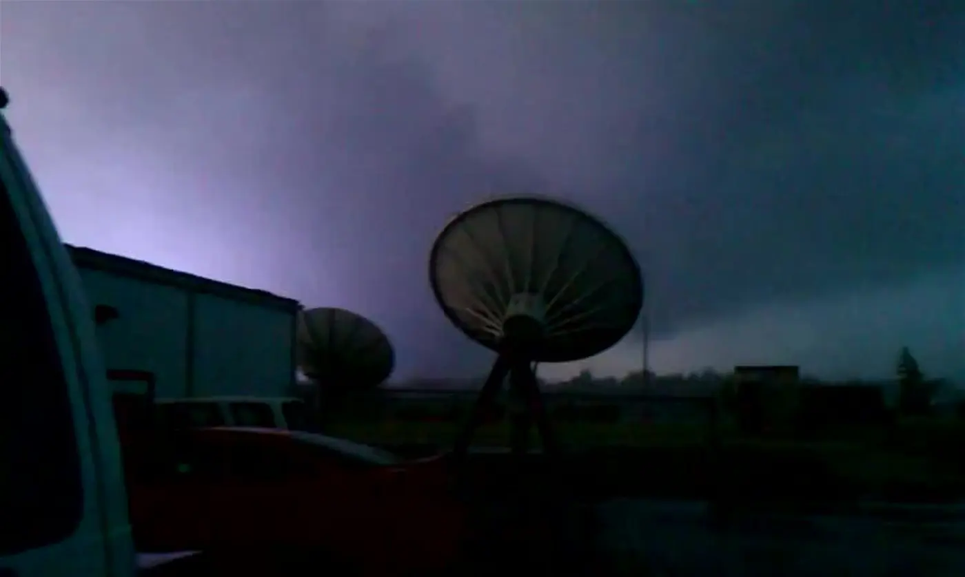 A dark, rain-wrapped tornado filling the frame, lit from below by the blue-white glare of an electrical flash.