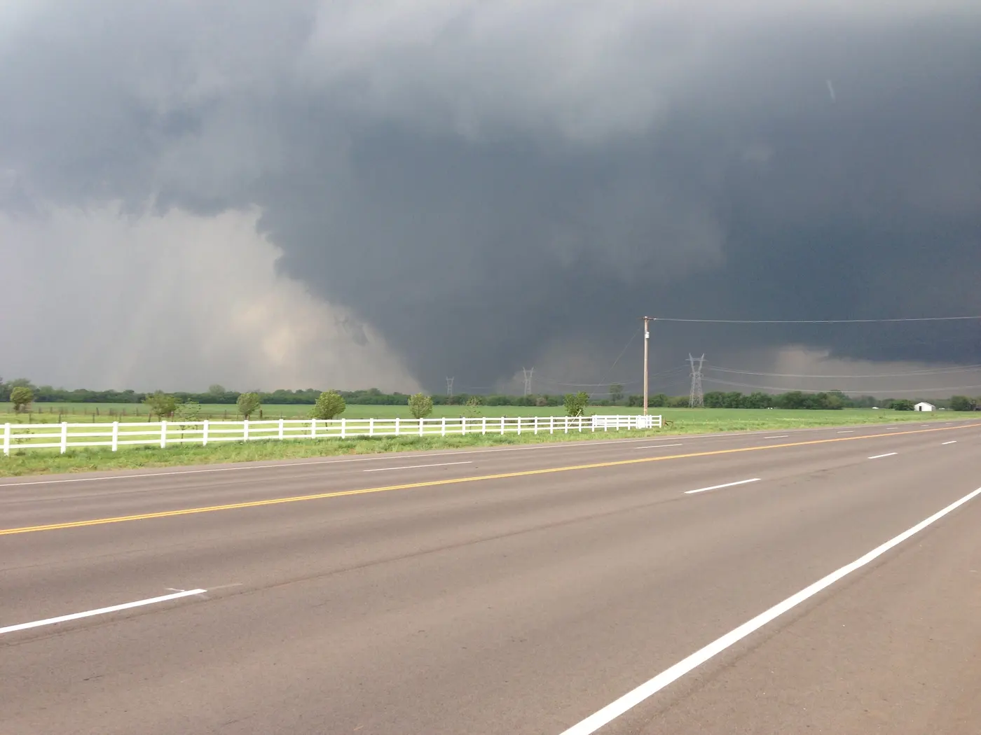 A very wide dark wedge tornado crossing open country beyond a white ranch fence and a road.