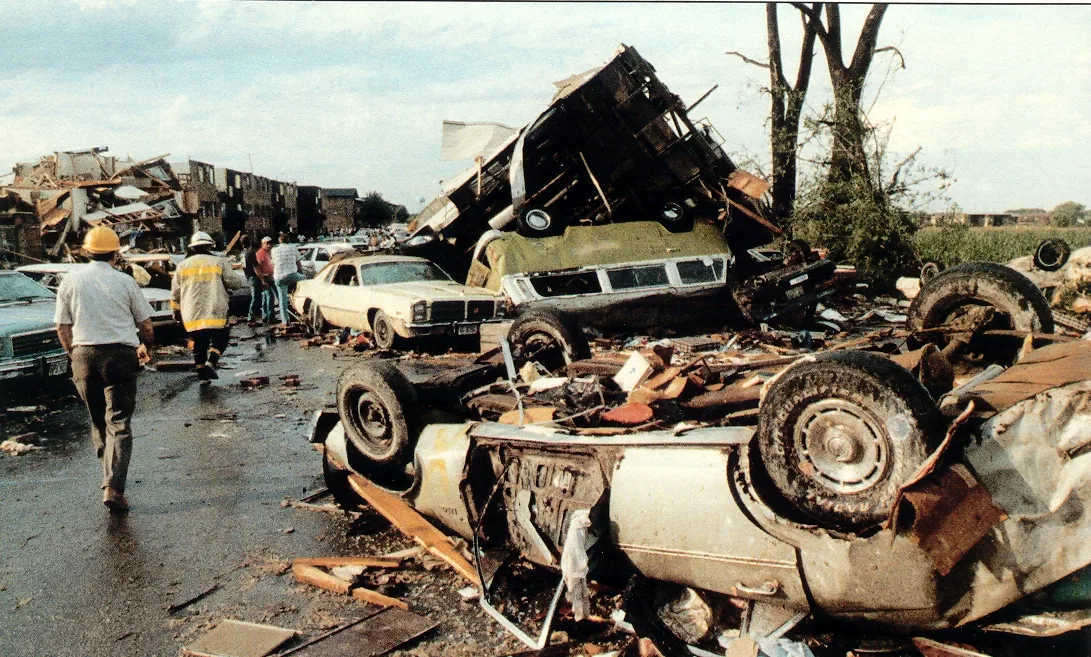 Overturned and crushed cars piled on a wet street, a firefighter and a man in a hard hat walking past, and a wrecked apartment building beyond.