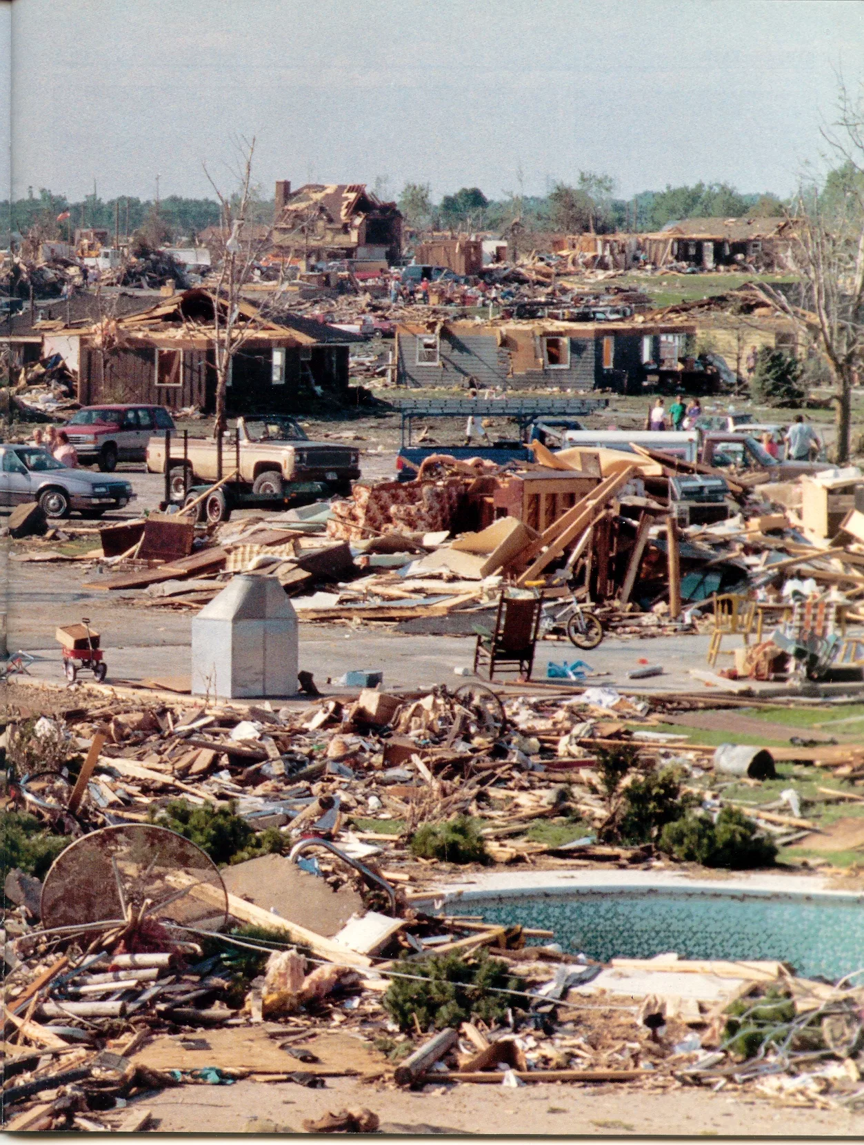 A levelled subdivision: splintered lumber, furniture and a chimney stack in the foreground beside a swimming pool, roofless houses and residents picking through debris beyond.