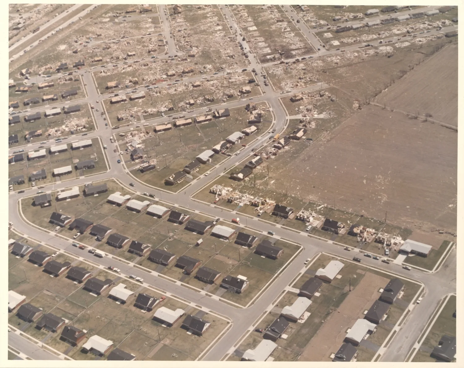 Aerial photograph of a suburban subdivision: rows of intact houses in the foreground give way to streets of bare slabs and scattered debris beyond a curving road.
