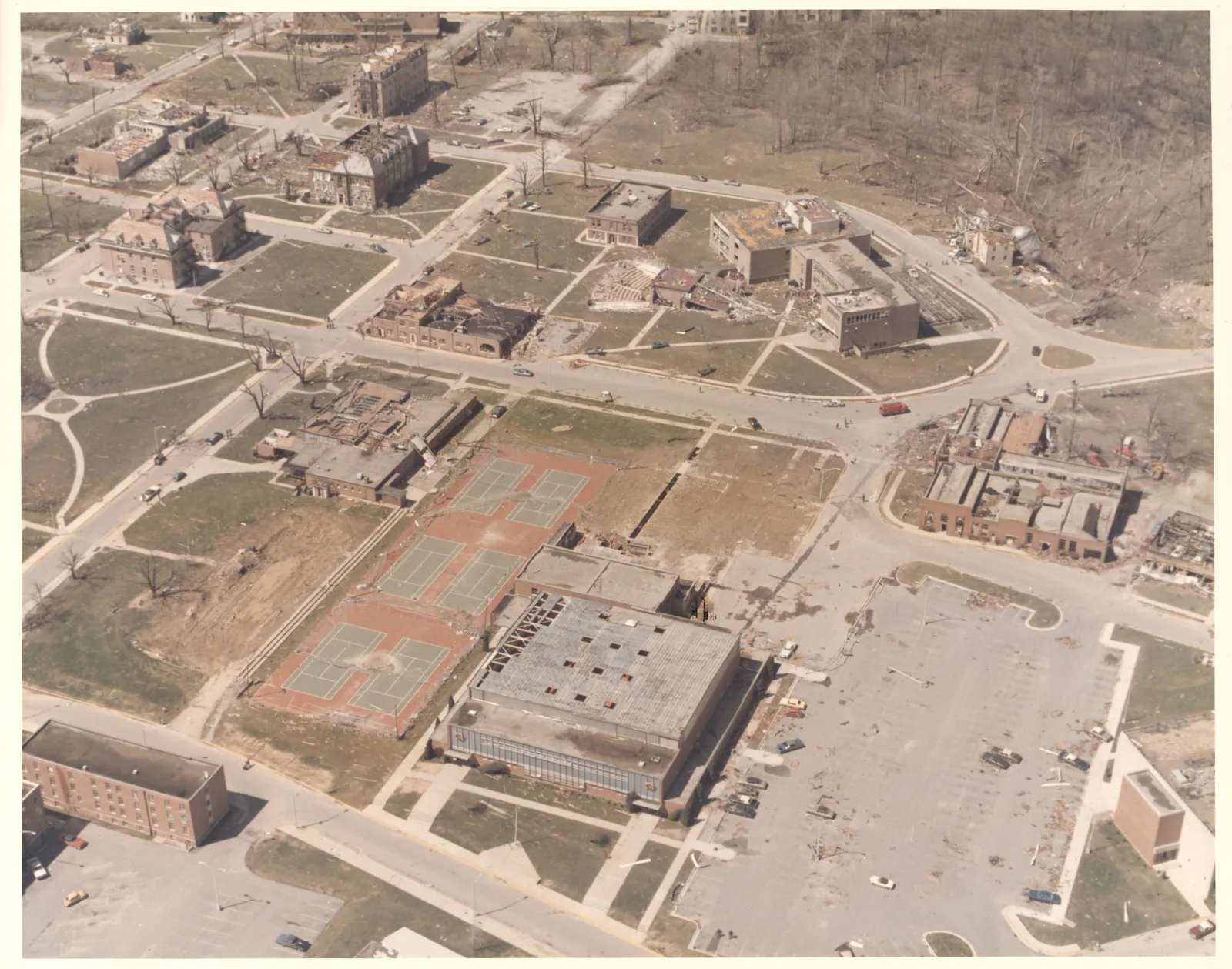 Aerial photograph of a university campus with damaged brick halls, a roofless gymnasium, torn-up tennis courts and a fallen water tower among stripped trees.