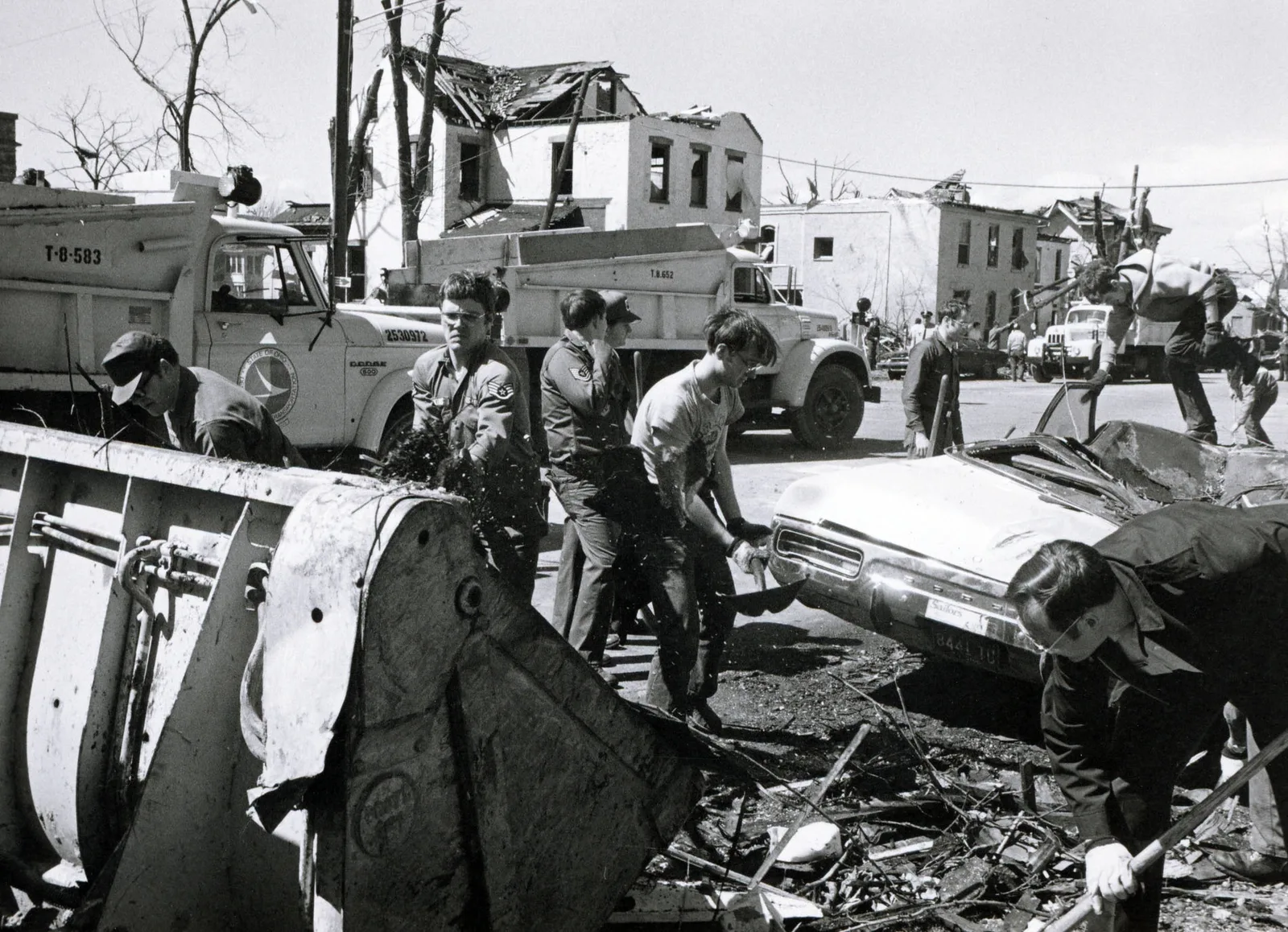 Black and white photograph of Ohio National Guard airmen and civilians clearing debris around a crushed car and a bulldozer blade, with roofless brick buildings and dump trucks behind.