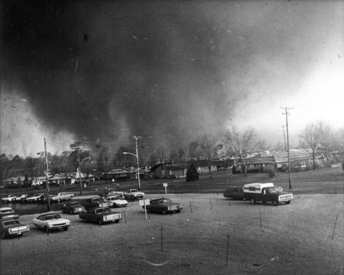 Black and white photograph of a wide, dark tornado with a lighter vortex on its left flank, looming over a row of single-storey houses beyond a car park.
